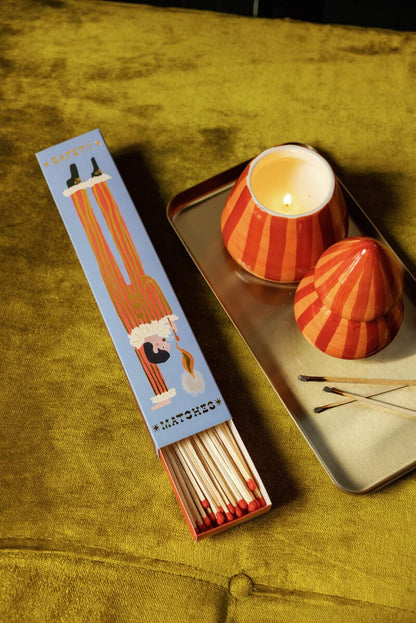 An open box of Jester - Extra Long Boxed Matches with a bright design sits on yellow next to a striped orange candle and tray, with burnt matches nearby for a playful decorative touch.
