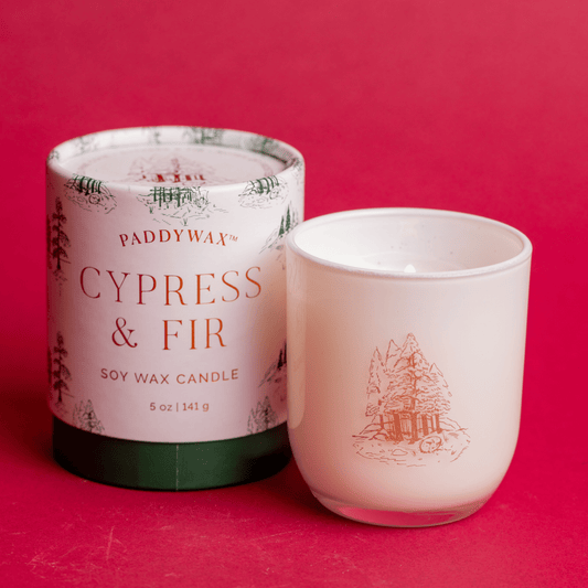 On red background, white opaque glass vessel with copper decal of fir trees; pictured next to white cylindrical candle holder with green fir trees on it