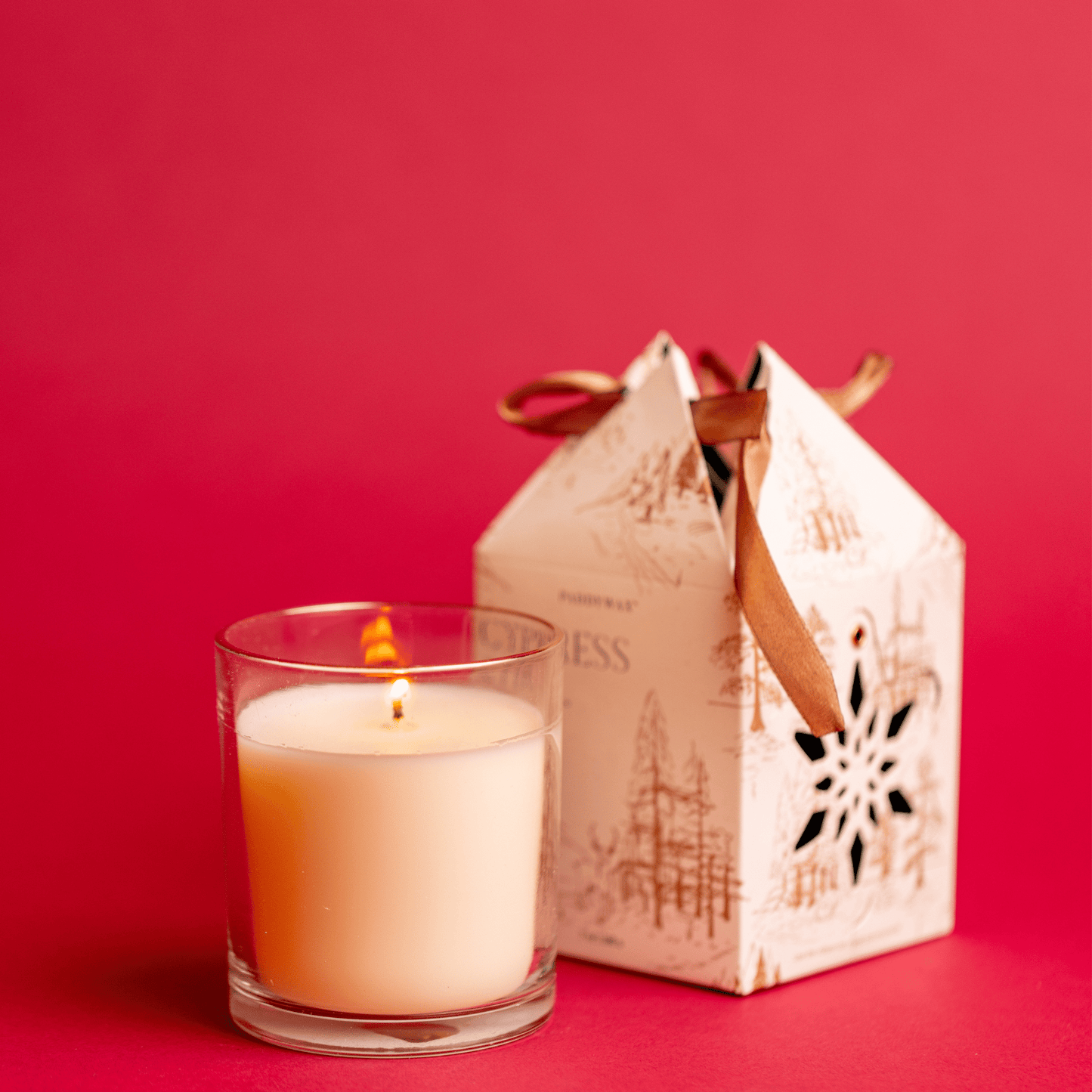 7 oz Glass Candle lit with Ornament Gift Box slightly out of focus on red background