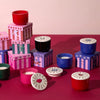 Colorful candles in red, green, blue, and black containers with illustrated lids and matching decorative boxes are arranged on a red and pink surface against a pale pink background.