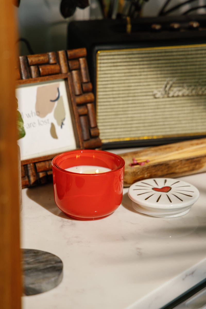 A lit Wild Fig & Rose - Charmed 8oz Candle in a glass jar rests on marble beside its ceramic lid, an abstract art frame, and a vintage-style speaker.