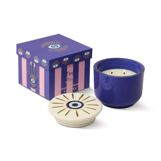 The Incense & Smoke - Charmed 8oz Candle, featuring a bold blue and white evil eye design, is displayed beside its ceramic lid and a purple-striped box with matching eye motifs.
