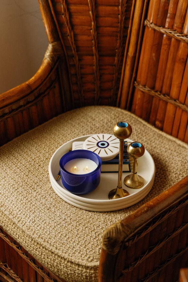 A white tray holding an Incense & Smoke - Charmed 8oz Candle, two brass candlesticks, and a closed book rests on a woven wicker chair.