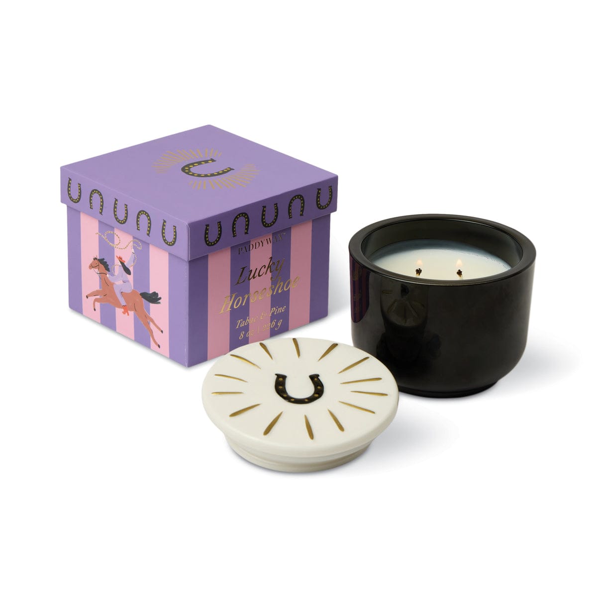 The Tabac & Pine - Charmed 8oz Candle, a black glass candle with ceramic lid featuring a horseshoe design, sits next to a purple box decorated with an illustration of a horse and rider.