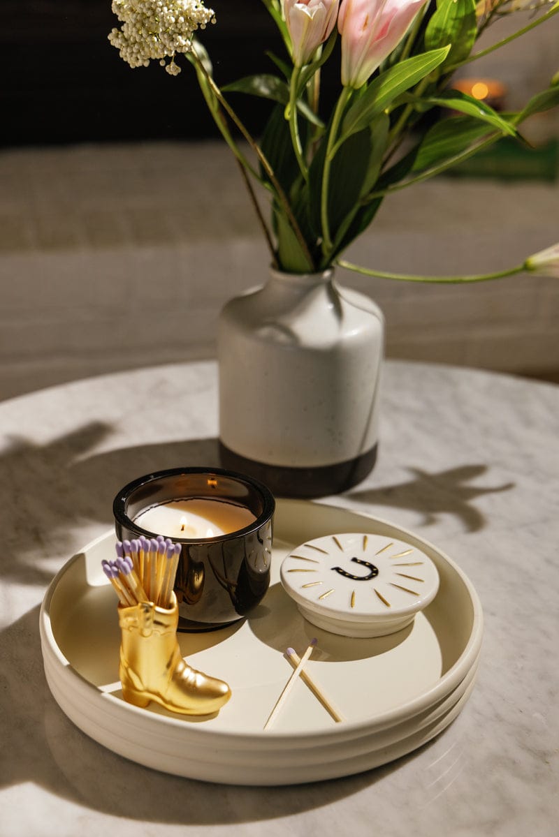 A lit Tabac & Pine - Charmed 8oz Candle, a decorative boot-shaped match holder with matches, and a flower-filled vase sit on a round tray atop a marble surface.
