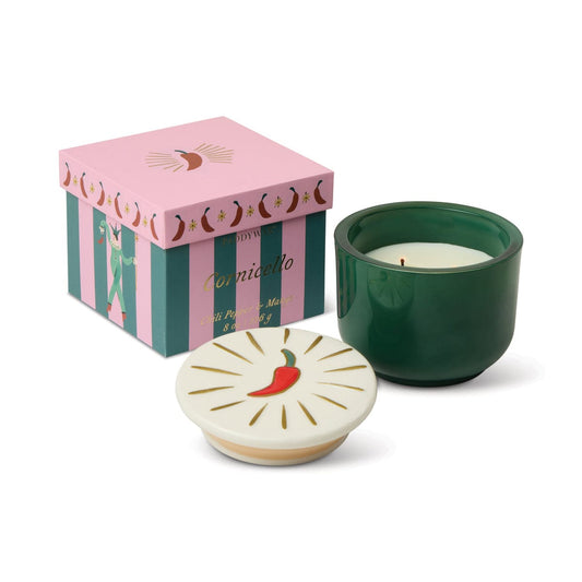 A Chili Pepper & Mango - Charmed 8oz Candle with a decorative ceramic lid featuring a red chili illustration sits beside a pink and green Cornicello-labeled box.