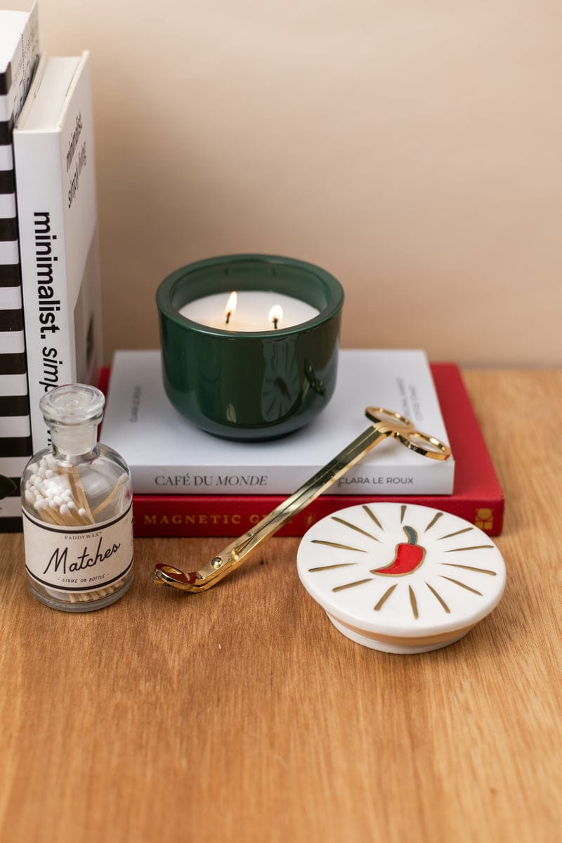 A Chili Pepper & Mango - Charmed 8oz Candle with a green ceramic lid, gold candle snuffer, glass jar of matches, dish with a red chili design, and stacked books are arranged on a wooden surface.