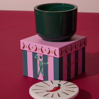 The Chili Pepper & Mango - Charmed 8oz Candle sits on a pink striped box featuring a circus performer; its white ceramic lid, decorated with a red chili pepper, is displayed in the foreground.