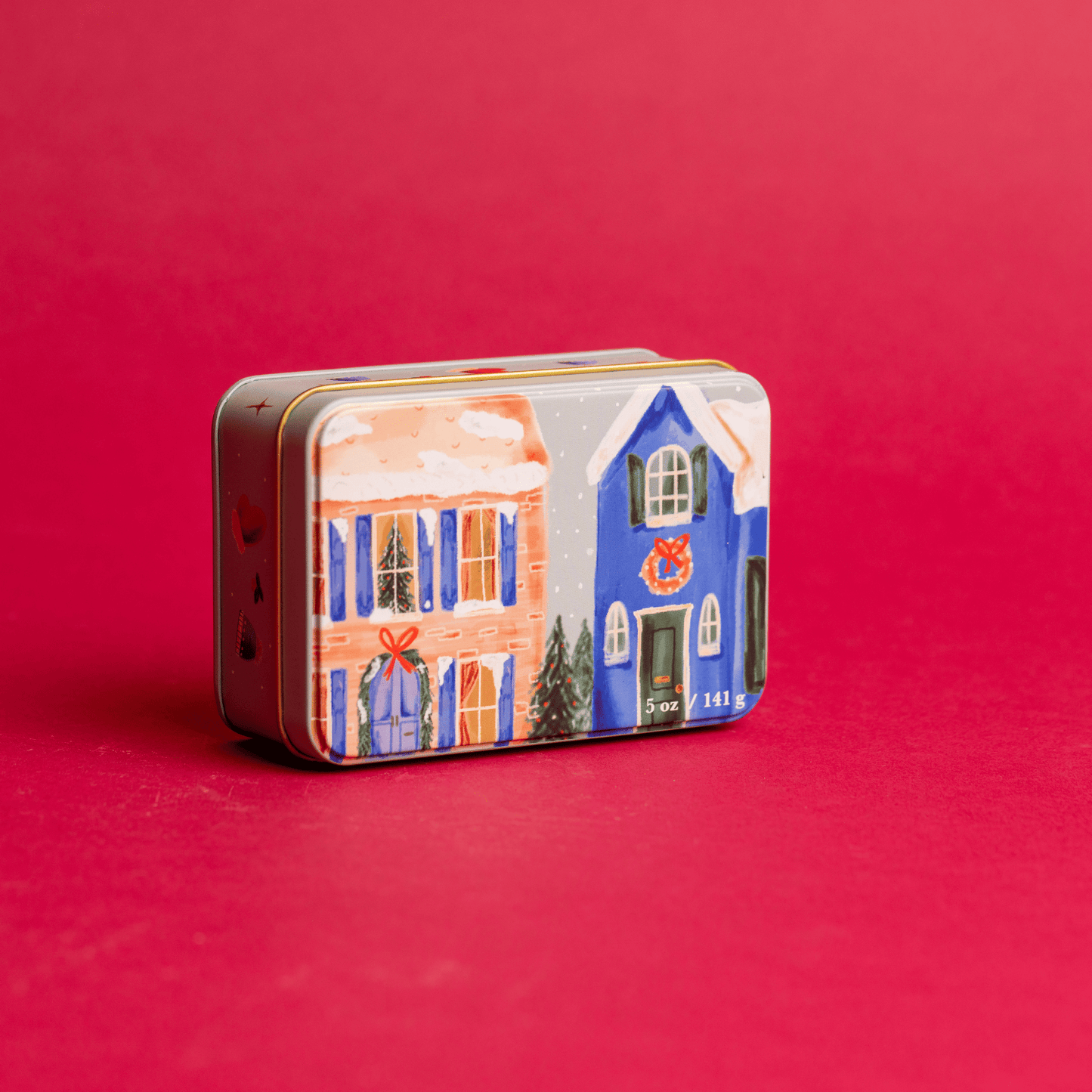 5 oz holiday tin with custom artwork; lid shows snow-covered houses decorated for the holidays; tin rests on a red background