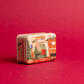 On a red background, 5 oz holiday tin with custom artwork; lid shows family room with piano and decorated tree 