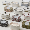 Assorted decorative candles in patterned, rectangular tins arranged on a white surface; some tins are open, showing lit candles inside.