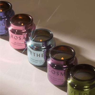 Five metallic candles in pink, blue, purple, and green jars are lit and arranged in a diagonal line on a light surface, with a lens flare across the image.