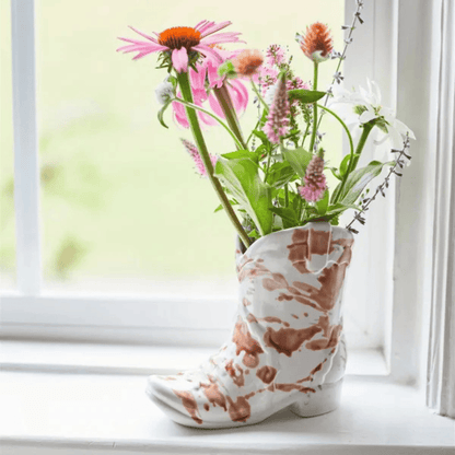 A Nashville Cowboy Boot - Desert Peach 6oz sits on a white windowsill, holding wildflowers with a window and greenery in the background, bringing a western-inspired touch and fragrant desert peach aroma to the scene.