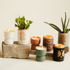 A collection of decorative candles and potted plants in modern ceramic holders, arranged on and beside natural stone slabs against a light background.