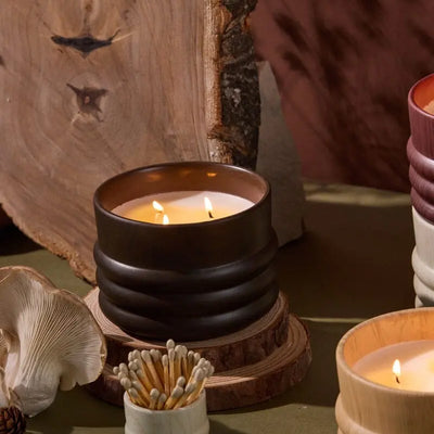 Three lit woodgrain candles in ribbed ceramic holders sit on a green surface near a White Washed Ceramic Wood Match Holder, a wooden log slice, and a mushroom.