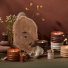 A display of lit candles in ribbed holders, stacked stones, wooden slab, matches, and dried flowers arranged on a green surface with a brown backdrop.