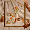 A gold picture frame is held in front of candles, flowers in vases, a pear, boxes, and reed diffusers, all arranged on a draped beige fabric background.