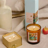 A lit amber glass candle and a lit Degas French Linen + Orris - Impressionist 3.5 oz Tin Candle sit on a beige cloth near a box, vase, orange, and apple. Both candles and the box have art-inspired fragrances with decorative artistic labels.