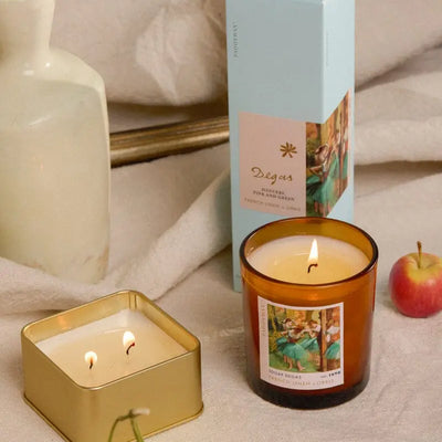 A lit amber glass candle and a lit Degas French Linen + Orris - Impressionist 3.5 oz Tin Candle sit on a beige cloth near a box, vase, orange, and apple. Both candles and the box have art-inspired fragrances with decorative artistic labels.