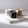 Three lit candles in black, white, and beige holders with minimalist hand illustrations, placed on a white stone surface with wooden matches nearby.