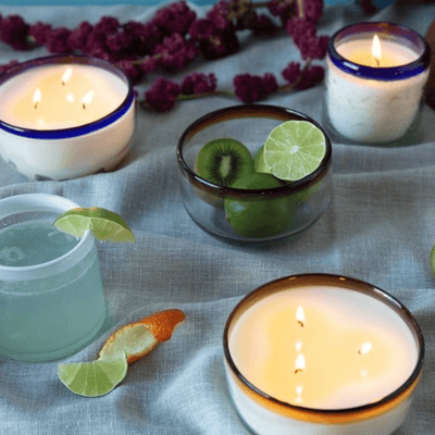 Lit candles in glass bowls, a drink garnished with lime, a glass with kiwi and lime slices, citrus peels, and purple flowers arranged on a blue fabric surface.