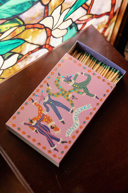 A box of Jesters - Large Rectangle Boxed Matches with colorful animal designs rests on a wooden surface near a stained glass window, bringing playful décor to the space.