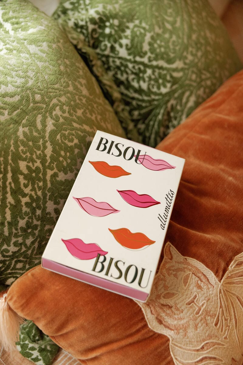 The Bisou Bisou - Large Rectangle Boxed Matches, featuring pink and orange lip art, rests on patterned pillows—a perfect Valentine’s gift.