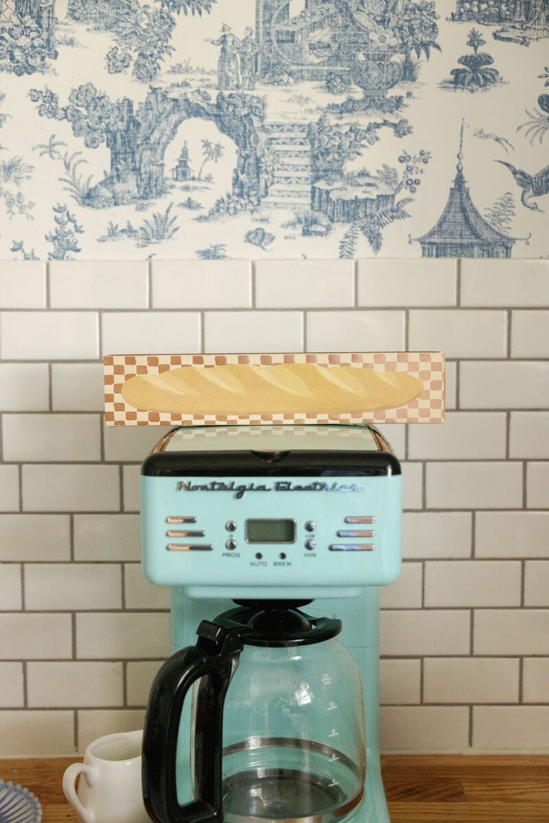 A blue Nostalgia Electrics coffee maker sits on a counter with an empty pot, while Baguette - Extra Long Boxed Matches in a checker-patterned box rest on top, against white subway tile and blue toile wallpaper.