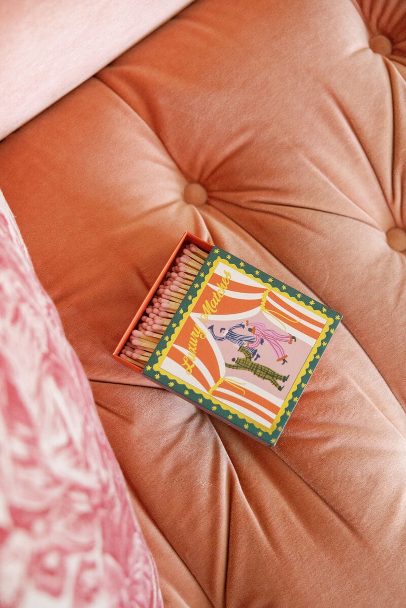 A box of Dancing Jesters - Square Boxed Matches with pink tips is open on a plush peach cushion beside a patterned pillow—a whimsical gift for any stylish space.