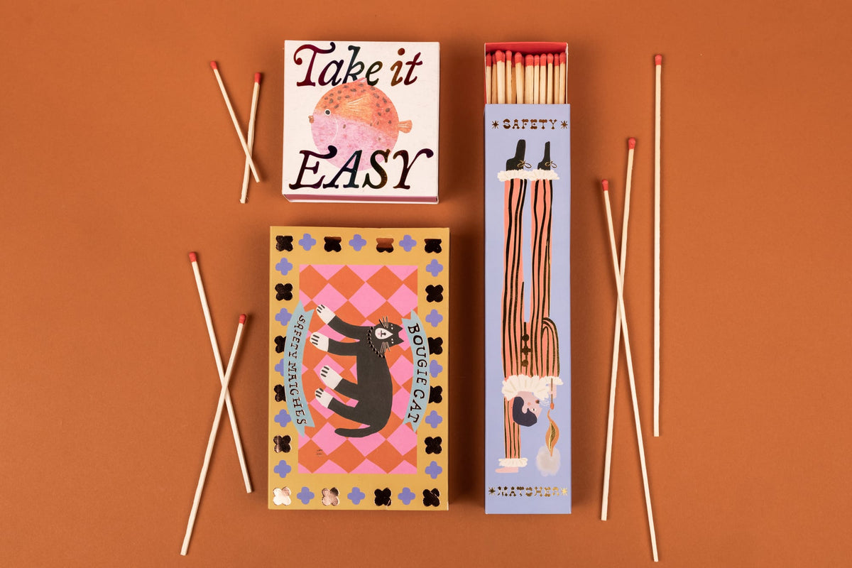 Three Jester - Extra Long Boxed Matches boxes with whimsical illustrations and scattered extra-long safety matches on an orange background, perfect as a gift for theater lovers.