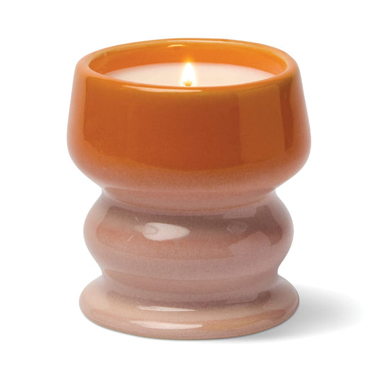A lit Infrared Mango - Melt 8.5 oz Candle, presented in a glossy, hand-glazed ceramic vessel featuring an orange upper section and a light pinkish base, emits a tropical fragrance and is photographed on a white background.