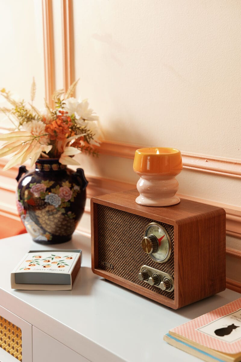 A wooden vintage radio sits on a white table beside a vase of flowers, a yellow candle in a holder, a floral box, and Oranges - Square Boxed Matches.