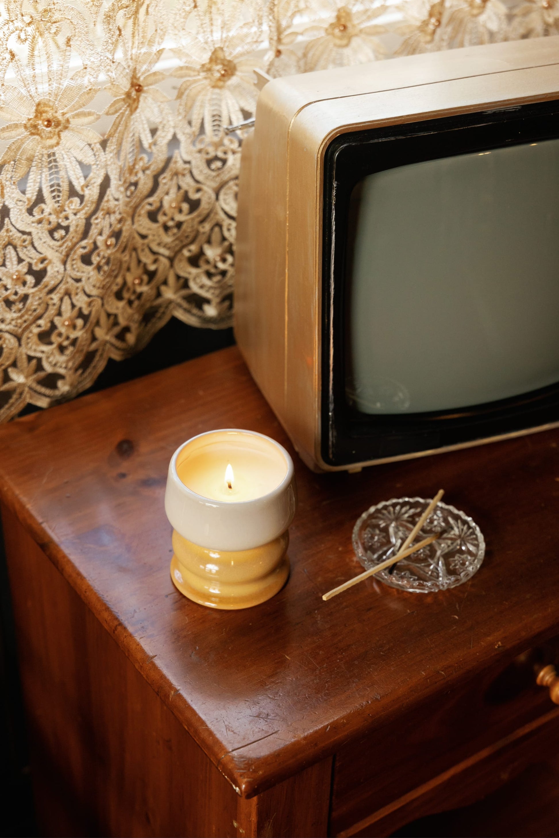 A Smoked Pistachio - Melt 8.5 oz Candle, an ashtray with a used match, and a vintage TV sit on a wooden table near a lace curtain, filling the air with a warm gourmand fragrance.