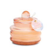 The Violet Vanilla - Beam 3 oz Candle is a round, peach-colored glass vessel with a lid and tag—ideal as a springtime fragrance candle or thoughtful gift.