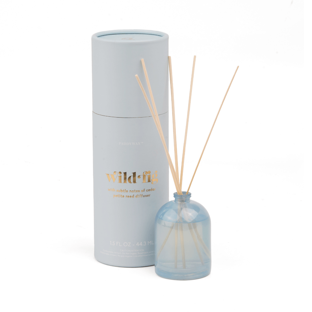 A pale blue milky glass Wild Fig - 1.5 oz Diffuser with wooden reeds sits next to a matching cylinder labeled in gold, offering a refreshing wild fig scent; further details appear on the container.