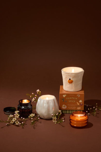 Four lit Pumpkin Spice - Beam 3 oz Candles in ceramic and glass containers—perfect as a holiday gift—are arranged on a brown surface with small flowers. One white Pumpkin Spice - Beam 3 oz Candle sits atop its box, while the other mini candles are placed around it in different holders.