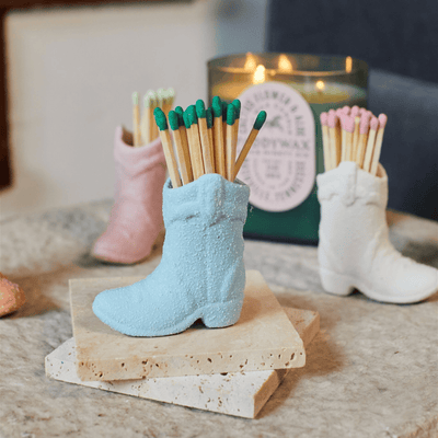 The Cowboy Boot Match Holder - Light Blue, a small ceramic holder in a pastel shade, rests on stone. A lit candle and dark chair add to its Western charm—perfect as a Nashville gift or home accent.