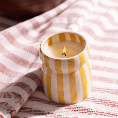 The Gold Coast - Cabana 6.5 oz Candle, in a yellow and white striped ceramic vessel, sits on a red and white striped cloth, infusing your space with fresh coastal scents.
