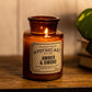 Apothecary Amber and Smoke candle on a wood shelf next to a plant
