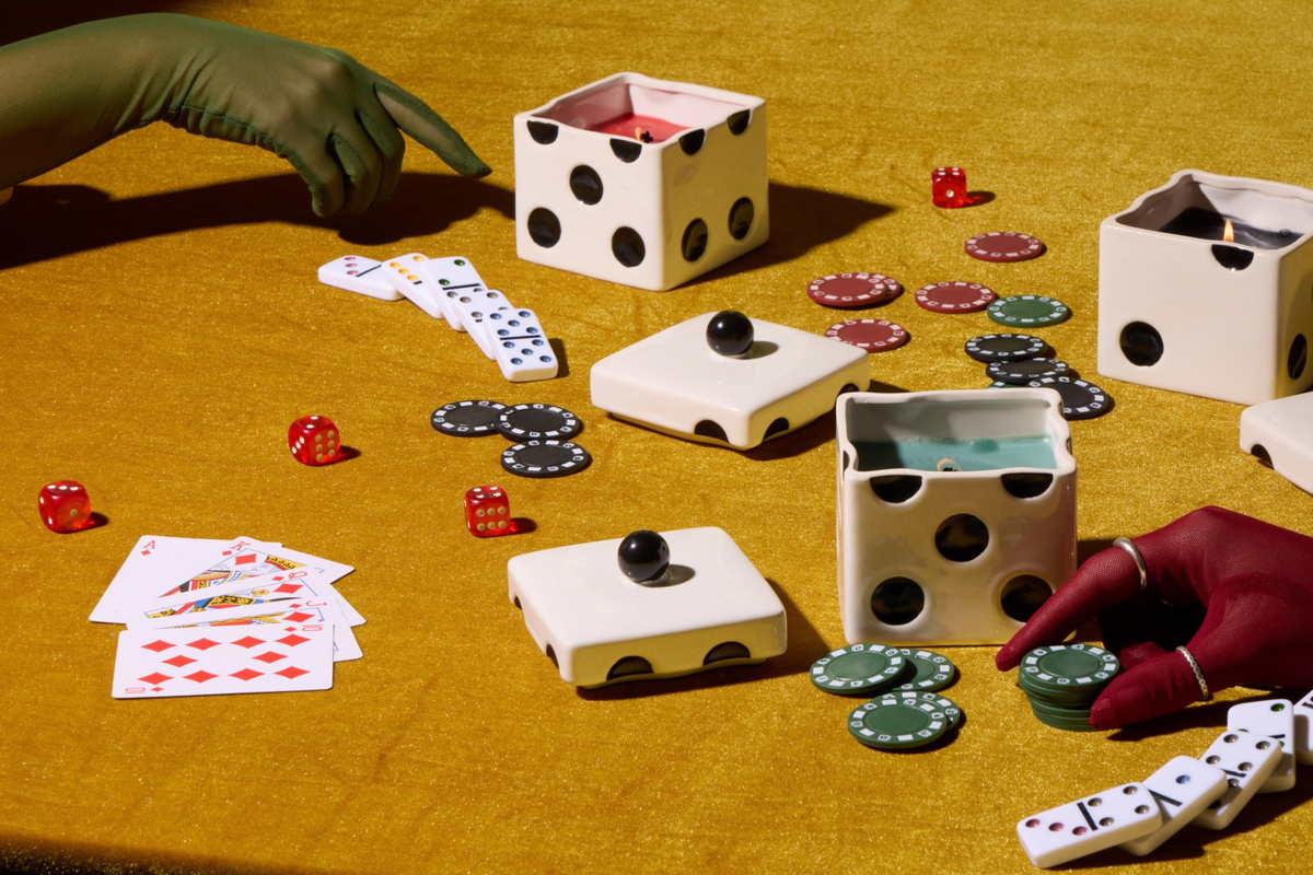 Hands in sheer green and red gloves interact with dice-shaped candles, playing cards, dominoes, and poker chips on a yellow velvet surface.