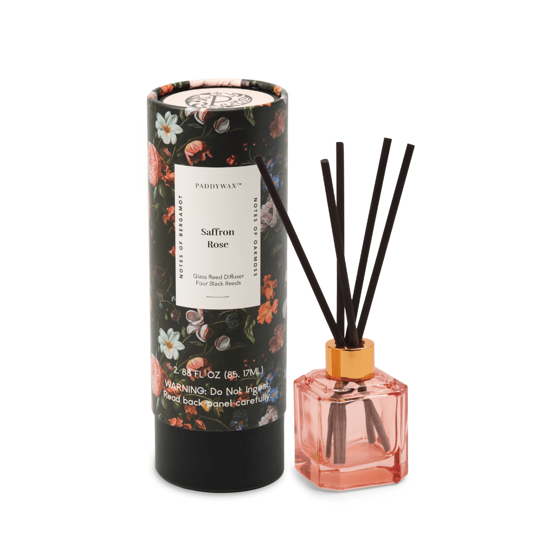 A floral-patterned cylindrical box labeled "Saffron Rose - Botanica Diffuser 2.88 oz" sits beside a glass reed diffuser filled with pink liquid and black reeds, both showcasing the elegant design of Paddywax’s Botanica Collection.