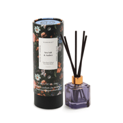 A floral-patterned cylindrical box labeled Sea Salt + Amber stands beside a purple glass reed diffuser with four black reeds and a gold collar, part of the Botanica Diffuser 2.88 oz collection.