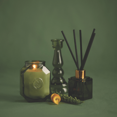 A lit green candle, a glass vase, the Wild Neroli - Botanica Diffuser 2.88 oz in black, and a lemon twist are arranged on a green surface against a matching green background.