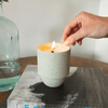 A hand lights a candle with a match. The candle is in a speckled ceramic holder and is placed on a book next to a glass bottle filled with water.