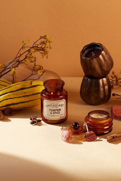 A brown apothecary jar labeled Pumpkin & Spice surrounded by a striped gourd, dried flowers, a small candle, and a stacked bronze candle holder on a beige surface.