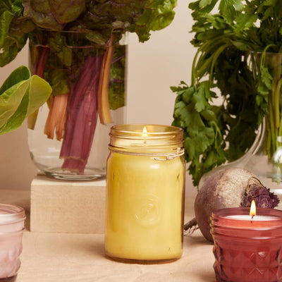 A lit yellow candle in a glass jar sits on a beige surface, surrounded by leafy greens, a beet, and two other colored candles.