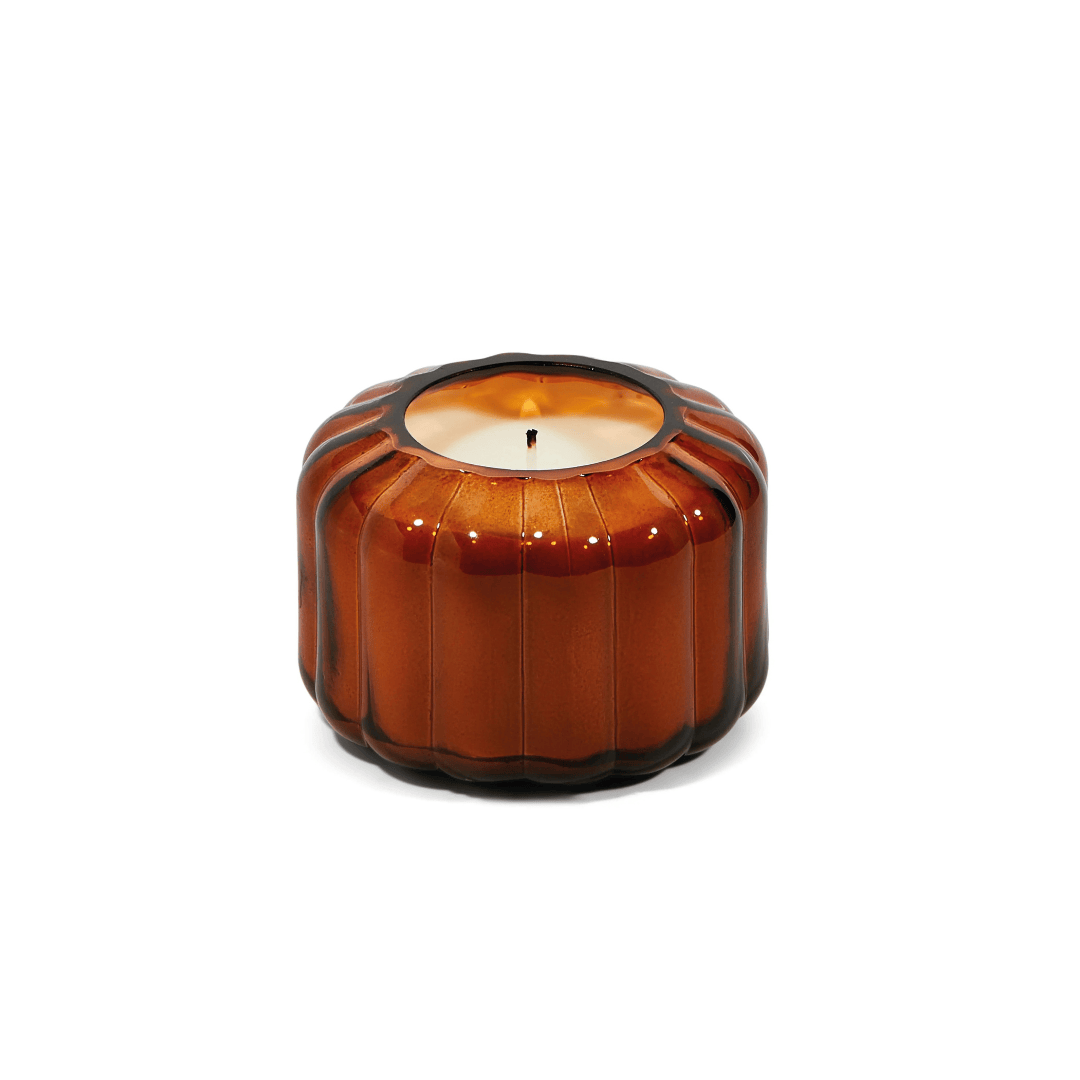The Tobacco Patchouli - Ripple 4.5 oz Candle, in a round ribbed glass container inspired by vintage vessels, glows warmly against a white background.