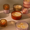 Several lit candles in ribbed glass containers of various pastel colors are arranged on a smooth surface, casting soft shadows.