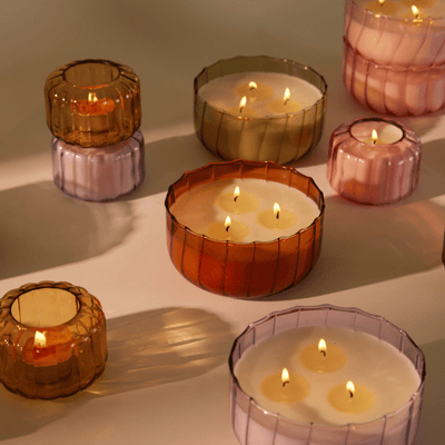 Several lit candles in ribbed glass containers of various pastel colors are arranged on a smooth surface, casting soft shadows.
