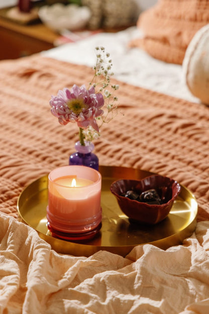 A lit Saffron Rose - Stack 7 oz Candle in a glass container, a small purple vase with pink flowers, and a red bowl rest on a gold tray placed over a textured orange blanket on the bed.